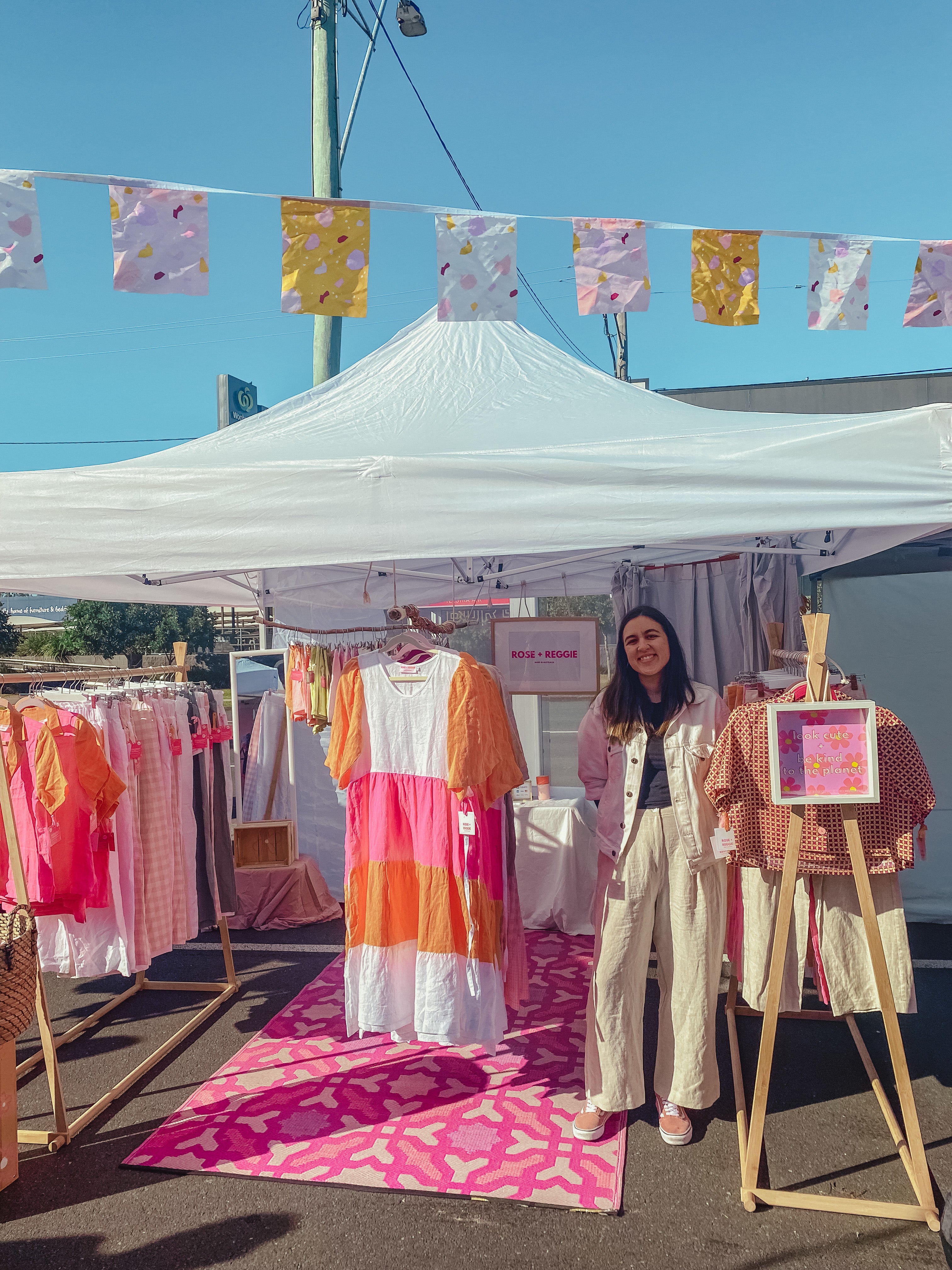 Woman standing in front of a clothing stall with colorful dresses and bunting at an outdoor market.